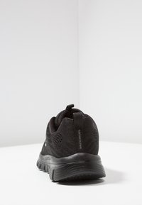 Svart Skechers träningsskon med mesh-ovandel och dämpad sula, sedd bakifrån på en vit yta och ljusgrå bakgrund.