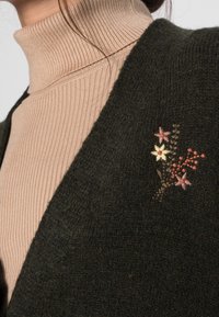 Primo piano di un maglione beige a collo alto a coste indossato sotto un cardigan scuro a maglia con piccoli fiori ricamati in rosa, giallo e arancione sul petto.