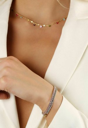 Guldhalsband med flerfärgade cirkulära berlocker och ett glittrande armband med växelvis rosa och klara stenar, buren på ett naket handled.