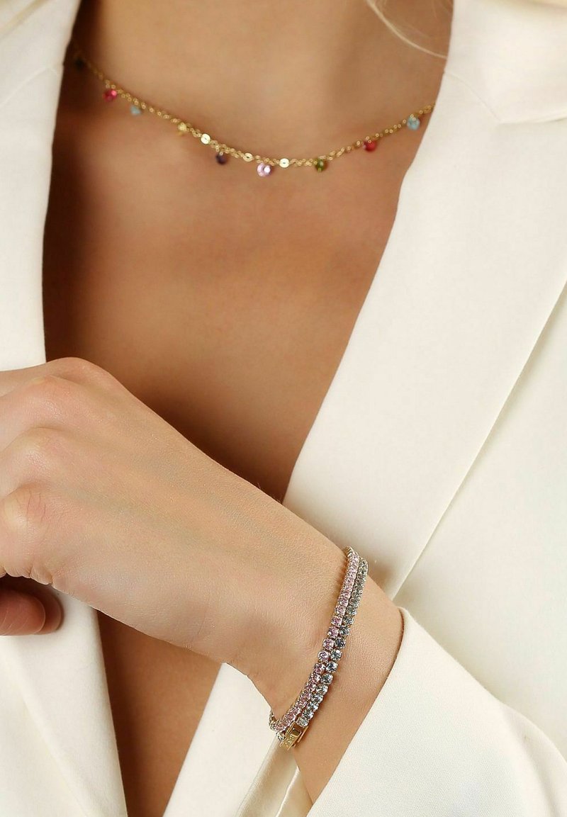 Guldhalsband med flerfärgade cirkulära berlocker och ett glittrande armband med växelvis rosa och klara stenar, buren på ett naket handled.