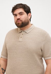 Polo beige texturé avec un col et une patte de boutonnage à trois boutons. Manches courtes avec bords côtelés.