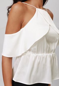 Haut blanc à épaules dénudées avec un design drapé, une texture plissée et une taille froncée. Tissu léger avec des bretelles fines.