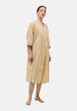 Femme debout pieds nus portant une robe midi beige ample avec des manches 3/4 et des poignets marron brodés sur un fond blanc uni.