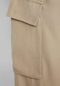 La tela beige presenta una superficie texturizada con un bolsillo prominente. Costuras limpias a lo largo de los bordes y un diseño sencillo perfilan la prenda.