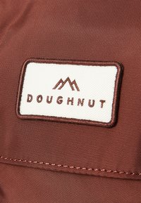 Brown fabric with a textured surface features a white rectangular patch. The patch displays the word "DOUGHNUT" and mountain logo in dark brown.