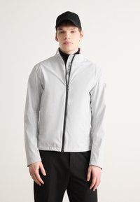 Ανοιχτό γκρι ζακέτα με φερμουάρ και ψηλό γιακά, με μαύρο φερμουάρ και λεπτομέρεια λογότυπου. Λείο υλικό, μακριά μανίκια και πλαϊνές τσέπες.