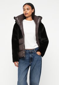 Mākslīgā kažokāda un quilted puffer jaka tumši brūnā un melnā krāsā, ar augstu apkakli, rāvējslēdzēju un sānu kabatām.
