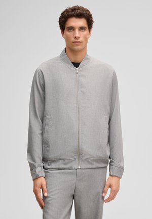 Jonge man draagt een lichtgrijze jas met rits en bijpassende broek, staand tegen een effen grijze achtergrond.