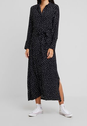 Femme portant une robe midi noire à manches longues, boutonnée, à pois blancs, avec une taille nouée, une fente latérale et des baskets blanches montantes.