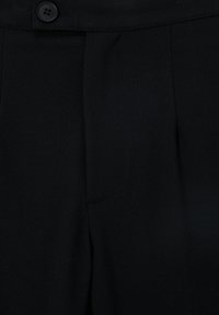 Gros plan d’un pantalon en tissu noir montrant une taille boutonnée et un détail de braguette cousue.