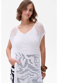 Witte gebreide tuniek met een groot open weefpatroon, korte mouwen en een V-halslijn, gecombineerd met zwarte met patronen versierde onderkleding. Soepele textuur, casual ontwerp.