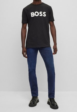 Muž v černém tričku s nápisem „BOSS“, tmavě modrých džínách a černých šněrovacích botách, stojící před jednoduchým světle šedým pozadím.