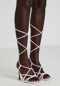 Sandali bianchi con tacco, dal design intrecciato, con cinturini incrociati che avvolgono la parte inferiore delle gambe e una punta quadrata.