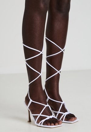 White heeled sandals with a strappy design, featuring crisscrossed straps that wrap around the lower legs and a square toe.