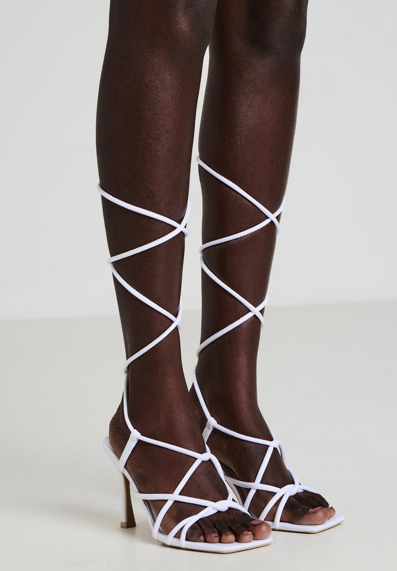 Sandali bianchi con tacco, dal design intrecciato, con cinturini incrociati che avvolgono la parte inferiore delle gambe e una punta quadrata.