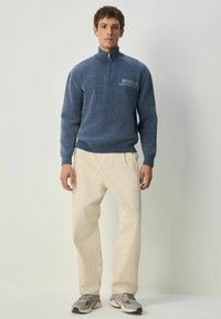 Tamsiai mėlynas vilnonis megztinis su ketvirtinės užtrauktuko apykakle ir už rašytą "BROOKLYN". Suderintas su šviesiai tan spalvos kelnėmis ir pilkais sportbačiais.