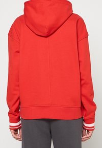 Personne vêtue d'un sweat à capuche rouge avec des poignets à rayures blanches et un pantalon gris foncé, vue de dos.