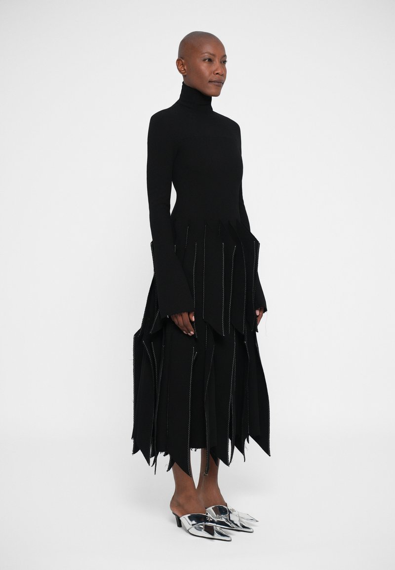 Black dress with a high neck, featuring jagged edges and intricate stitching. Paired with metallic, pointed-toe mules. Smooth texture.