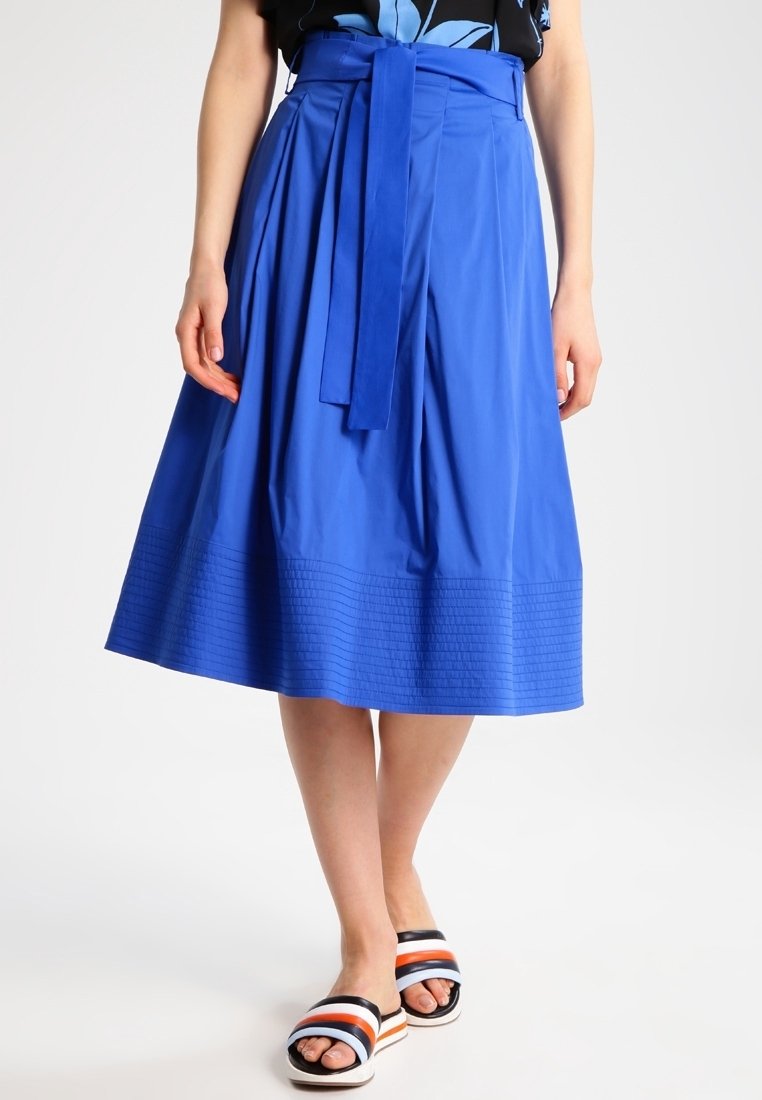 Femme portant une jupe bleu vif longueur genou avec une ceinture à la taille et des sandales à enfiler rayées à bouts ouverts sur fond blanc.