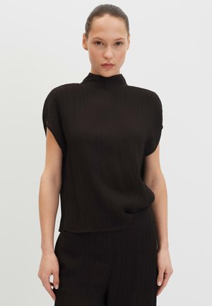 Femme aux cheveux tirés en arrière portant un haut noir texturé à manches courtes et un pantalon assorti, debout devant un fond uni clair.