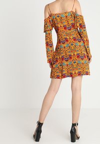 Vestido amarillo de hombros caídos con un patrón floral en azul y rojo, corsé ajustado, falda acampanada y mangas largas. Se lleva con botas de tobillo negras de tacón.