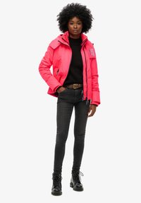 Jasnoróżowa, ocieplana kurtka z wysokim kołnierzem, z zamkiem z przodu i detalem z logo. Noszona na czarnym swetrze, w zestawieniu z ciemnymi dżinsami i czarnymi butami.