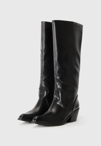 Bottes noires en cuir, arrivant au genou, avec un bout pointu et un talon épais. Texture lisse avec des détails de couture subtils sur les côtés.