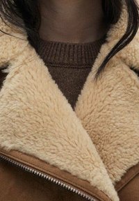 Veste en shearling marron avec un col en fausse fourrure beige, portée sur un pull marron foncé. Elle comporte une fermeture éclair, une texture douce et un aspect superposé.