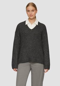 Kraftvoller schwarzer Strickpullover mit V-Ausschnitt und strukturierter Oberfläche. Kombiniert mit einem weißen Hemd mit Kragen darunter und grauen Hosen.