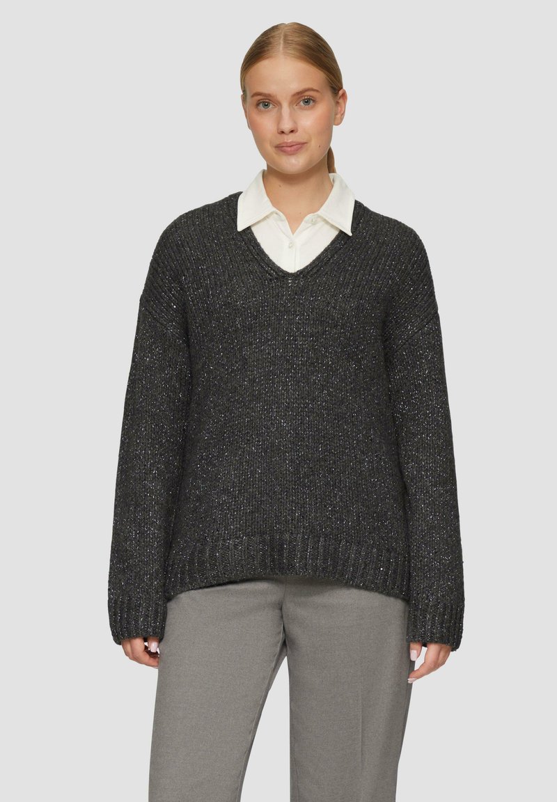 Kraftvoller schwarzer Strickpullover mit V-Ausschnitt und strukturierter Oberfläche. Kombiniert mit einem weißen Hemd mit Kragen darunter und grauen Hosen.