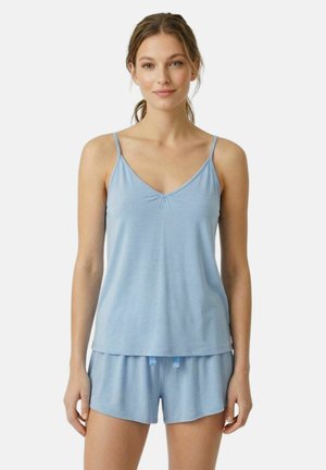 Jonge vrouw staat recht voor zich uit kijkend, draagt een lichtblauwe mouwloze top en bijpassende korte broek tegen een effen witte achtergrond.
