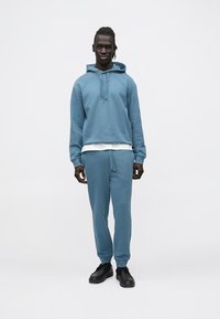 Blauwe hoodie en bijpassende joggingbroek gemaakt van zachte stof. De hoodie heeft een voorzak en verstelbare trekkoorden. Zwarte sneakers maken de look compleet.