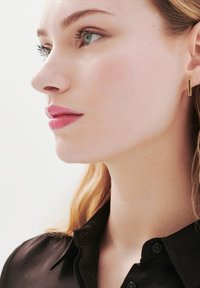 Złote geometryczne kolczyki w kształcie stud, gładka faktura, noszone na prawym uchu. Modelka ma jasnobrązowe włosy, ubrana w czarną satynową koszulę z guzikami.