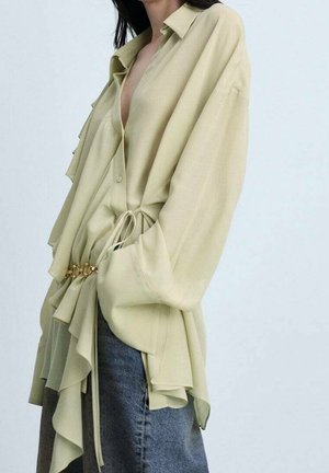 Femme portant un chemisier beige oversize avec des détails à volants et une ceinture chaîne dorée, assorti à un jean bleu à jambes larges.