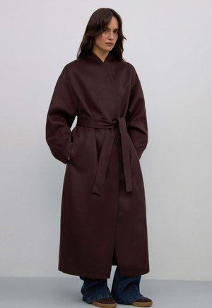 Long, dark brown coat made from textured fabric, featuring a wrap design, wide sleeves, a tie belt at the waist, and side pockets.