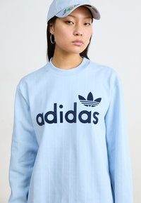 Sudadera azul clara con rayas verticales, que presenta un gran logotipo negro de "adidas" y un emblema de trébol en el pecho. Corte casual, escote redondo.