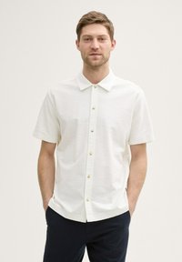 Wit shirt met korte mouwen en een gestructureerde afwerking, met een knoopsluiting aan de voorkant en lichtgekleurde knopen. Draagt het met donkere broeken, handen in de zakken.