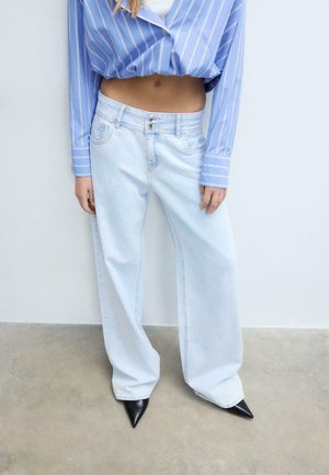 Osoba ubrana w jasnoniebieskie szerokie dżinsy, czarne spiczaste buty oraz niebieską paskowaną krótka koszulę, stojąca przy białej ścianie.