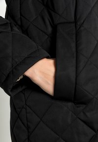 Handen inskjuten i sidofickan på en svart vadderad jacka som visar detaljerad sömnad och tygtextur.