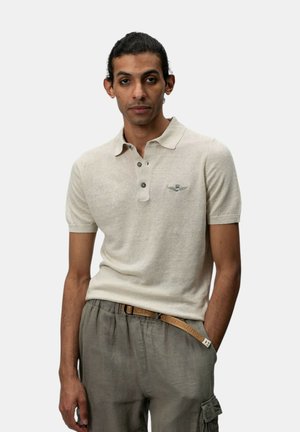 Uomo con capelli scuri legati dietro, indossa una polo beige a maniche corte e pantaloni grigi con cintura, in piedi davanti a uno sfondo bianco semplice.