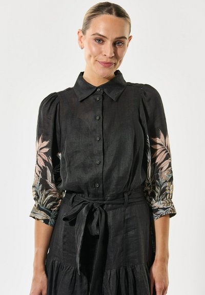 Robe chemise en lin noir avec un devant à boutons, des manches courtes bouffantes ornées de broderies florales et un détail de taille nouée.
