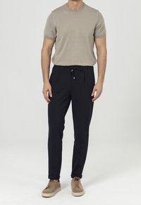 Mężczyzna ubrany w beżową koszulę z krótkim rękawem, czarne spodnie z troczkiem i beżowe sneakersy, stojący na tle białym.