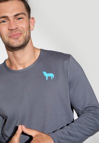 Długi rękaw szara koszulka z miękkiego materiału, z jasnoniebieskim sylwetką psa na klatce piersiowej. Projekt jest prosty i nowoczesny.