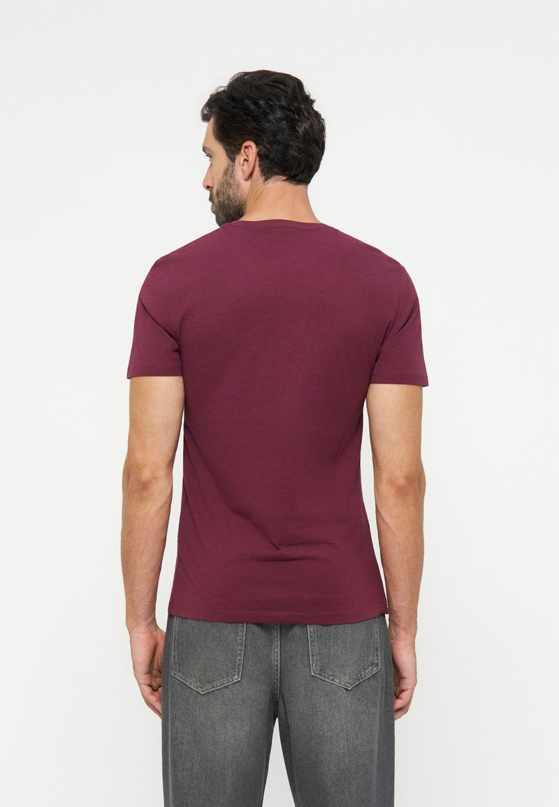 Man met donker haar die een effen bordeauxrode korte mouwen T-shirt en grijze spijkerbroek draagt, gezien van achter tegen een witte achtergrond.