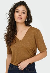 Bruine v-hals t-shirt gemaakt van zacht materiaal, korte mouwen, relaxte fit, gedragen met donkerblauwe jeans. Accessoires omvatten zilveren ringen.