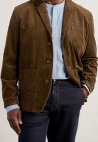 Brązowa marynarka z korduroju z kołnierzem z klapami, zapięciem na guziki, przednimi kieszeniami, noszona na jasnoniebieską koszulę i ciemne dżinsy.