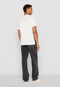 White short-sleeve t-shirt with a round neckline, paired with dark denim jeans and beige sneakers, viewed from the back.