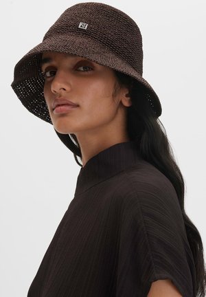 Frau mit langen dunklen Haaren trägt einen dunkelbraunen, strukturierten Bucket Hat und ein dunkles, hochgeschlossenes Oberteil, schaut leicht seitlich vor weißem Hintergrund.