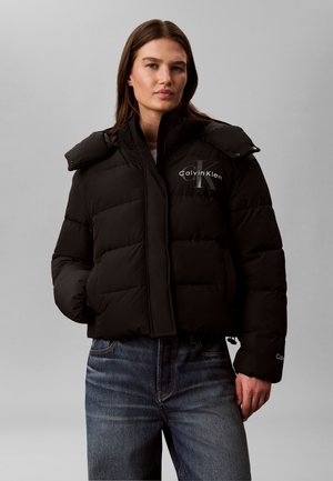 Kvinde iført en sort Calvin Klein dunjakke med logo på bryst og ærme, kombineret med blå jeans, stående mod en ensfarvet grå baggrund.