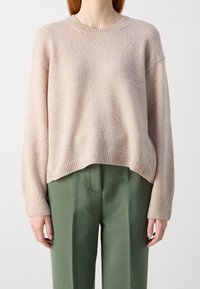 Pull rose clair avec un col rond et des épaules tombantes. Il présente des poignets et un ourlet côtelés, associé à un pantalon vert. Texture douce.